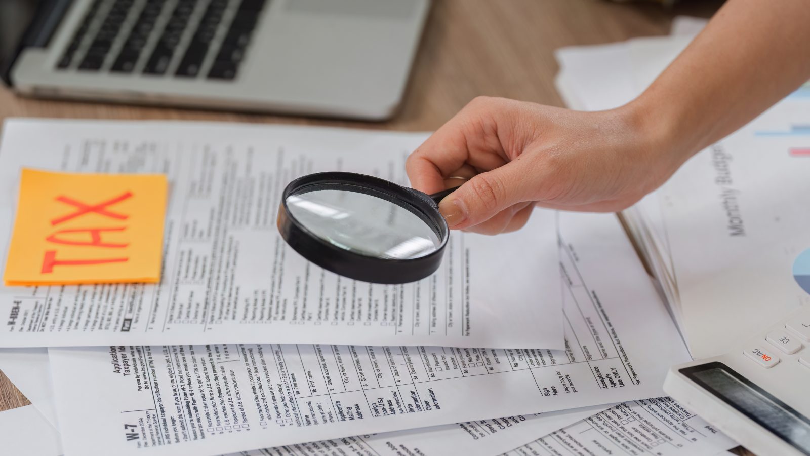 A person using a magnifying glass to scrutinize tax documents on a desk © Imagn Images via Vecteezy.com